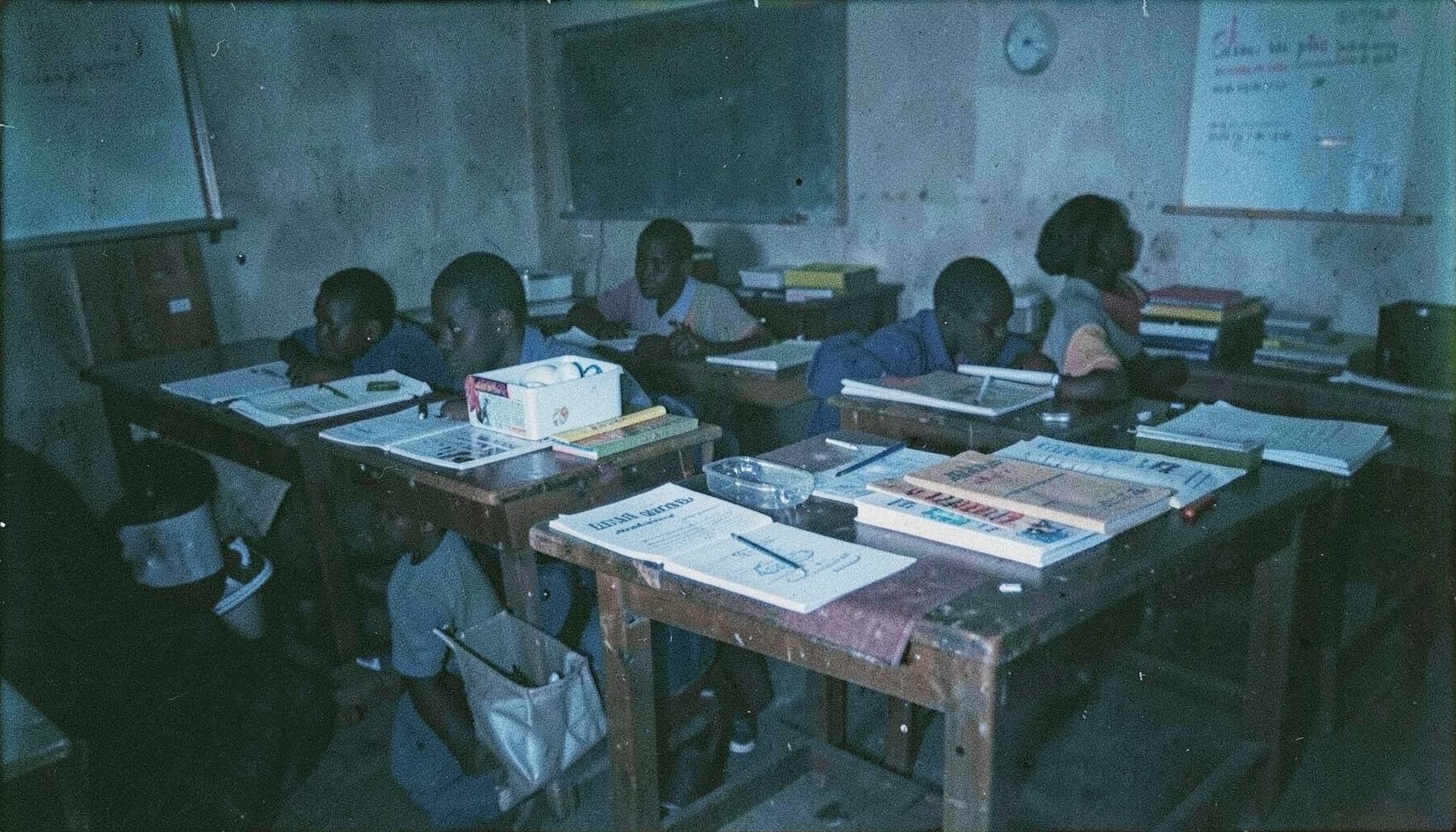 A dark-toned analog film photograph with visible grain showing a Ghanaian classroom scene with educational materials and learning equipment arranged on wooden tables