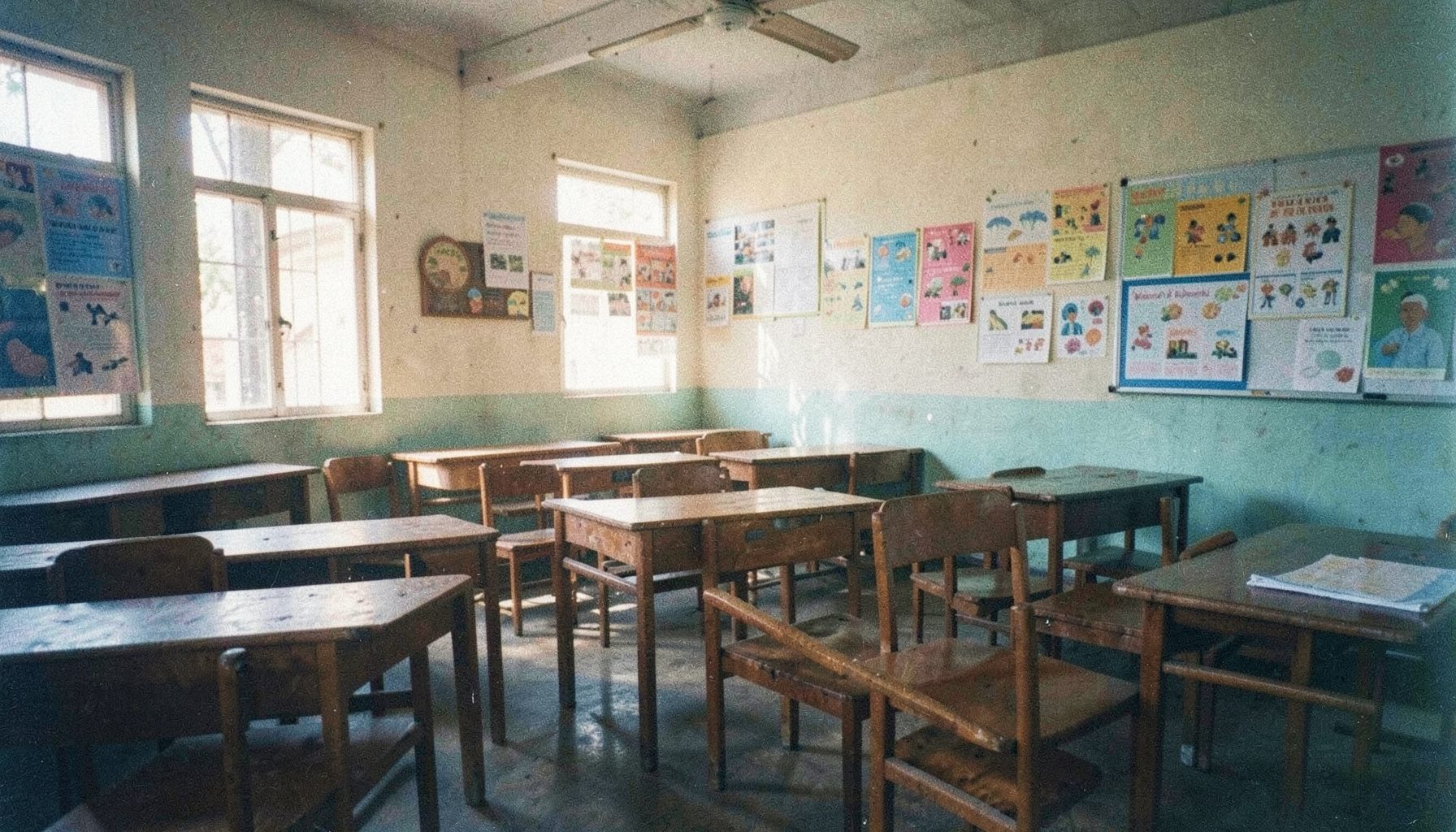 A warm analog film photograph with visible grain showing a sunlit Ghanaian primary classroom interior with wooden desks and colorful learning materials on the walls