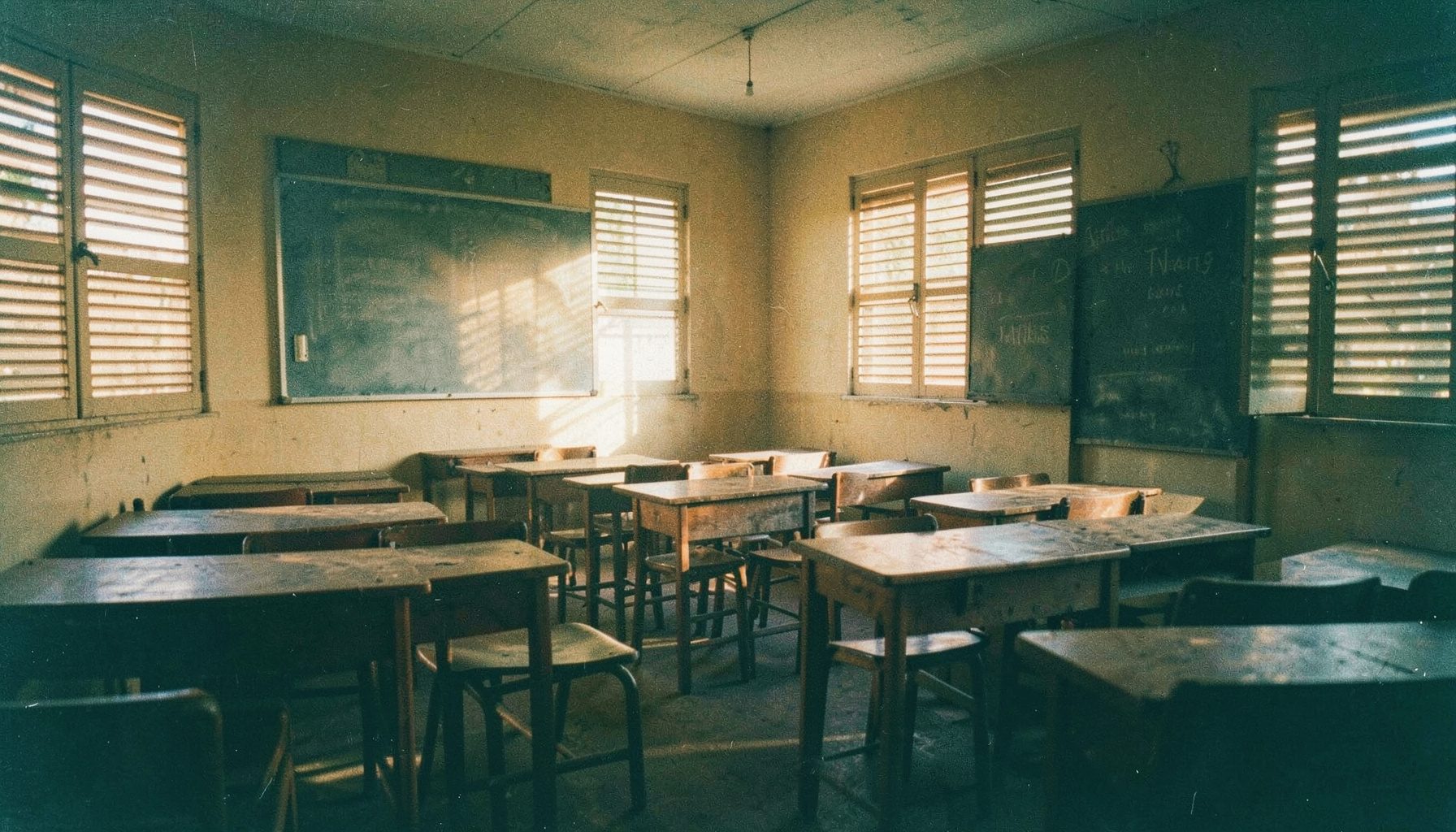 Wide landscape photograph of a quiet Ghanaian classroom with wooden desks and a chalkboard, warm golden afternoon light