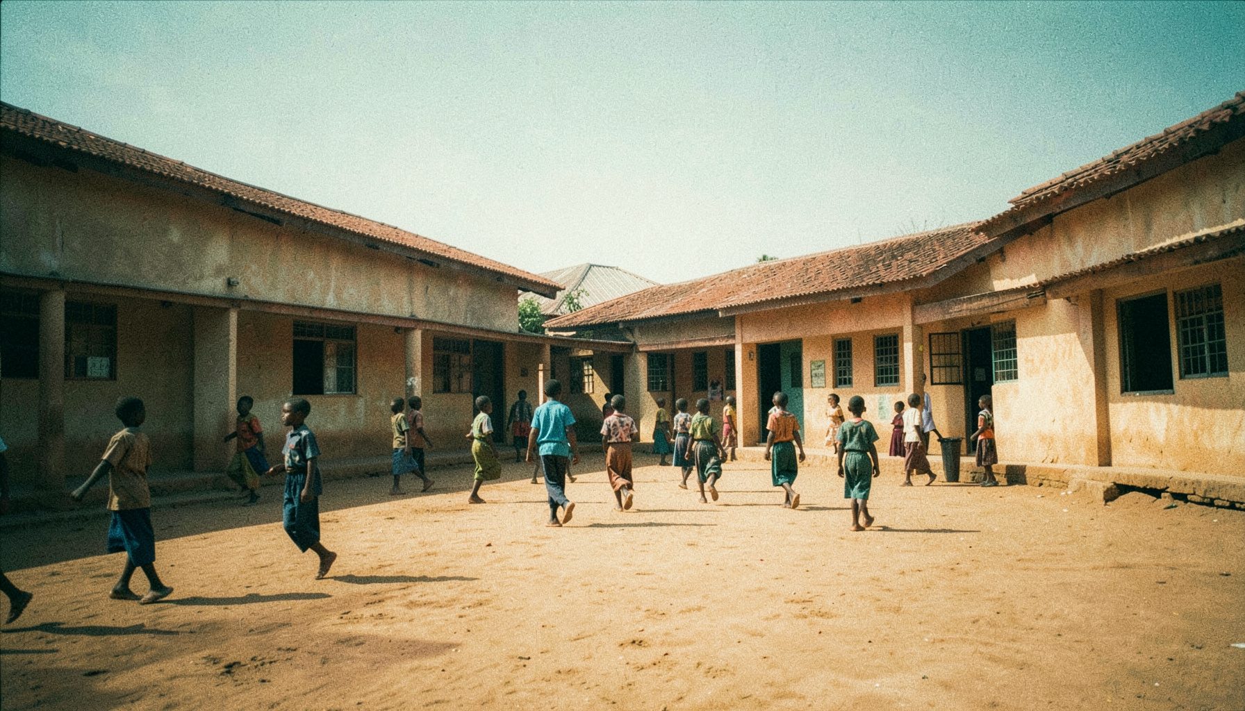 A wide-angle view of a rural Ghanaian primary school building with children in uniforms walking across a dusty courtyard under bright sunlight